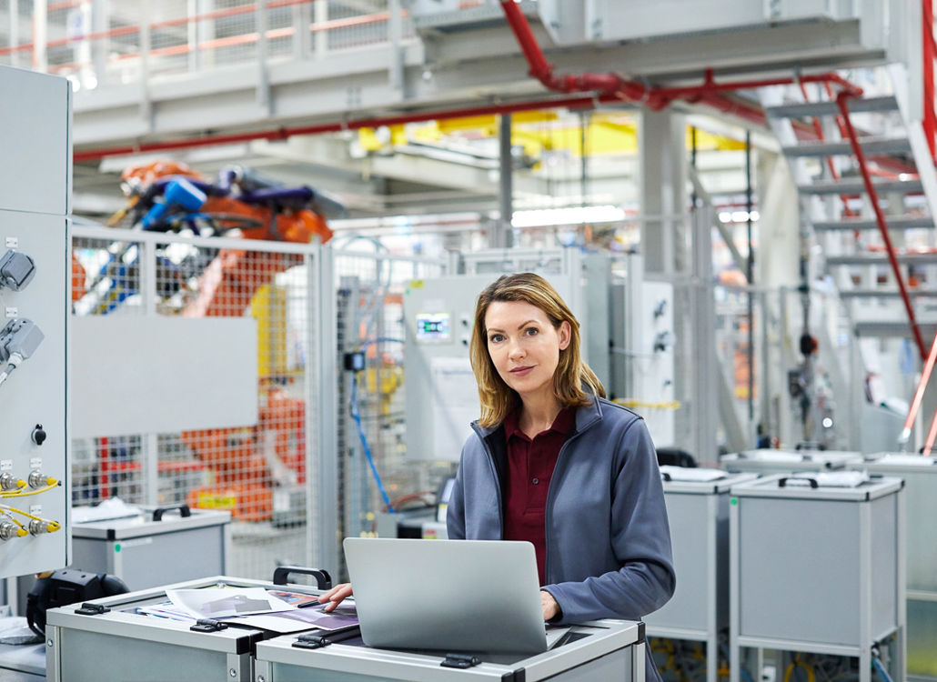 En selvsikker kvinnelig ingeniør jobber på en bærbar PC i en moderne industrifabrikk og overvåker automatiserte produksjonssystemer.
